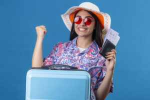 beautiful tourist woman in summer hat wearing red sunglasses holding travel suitcase and passport with tickets looking at camera with smile on face happy and positive raising fist after a victo